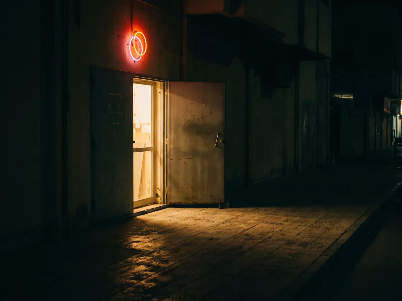The workshop door at night, half open, lamplight spilling onto the wet pavement, a small neon loop lit above it