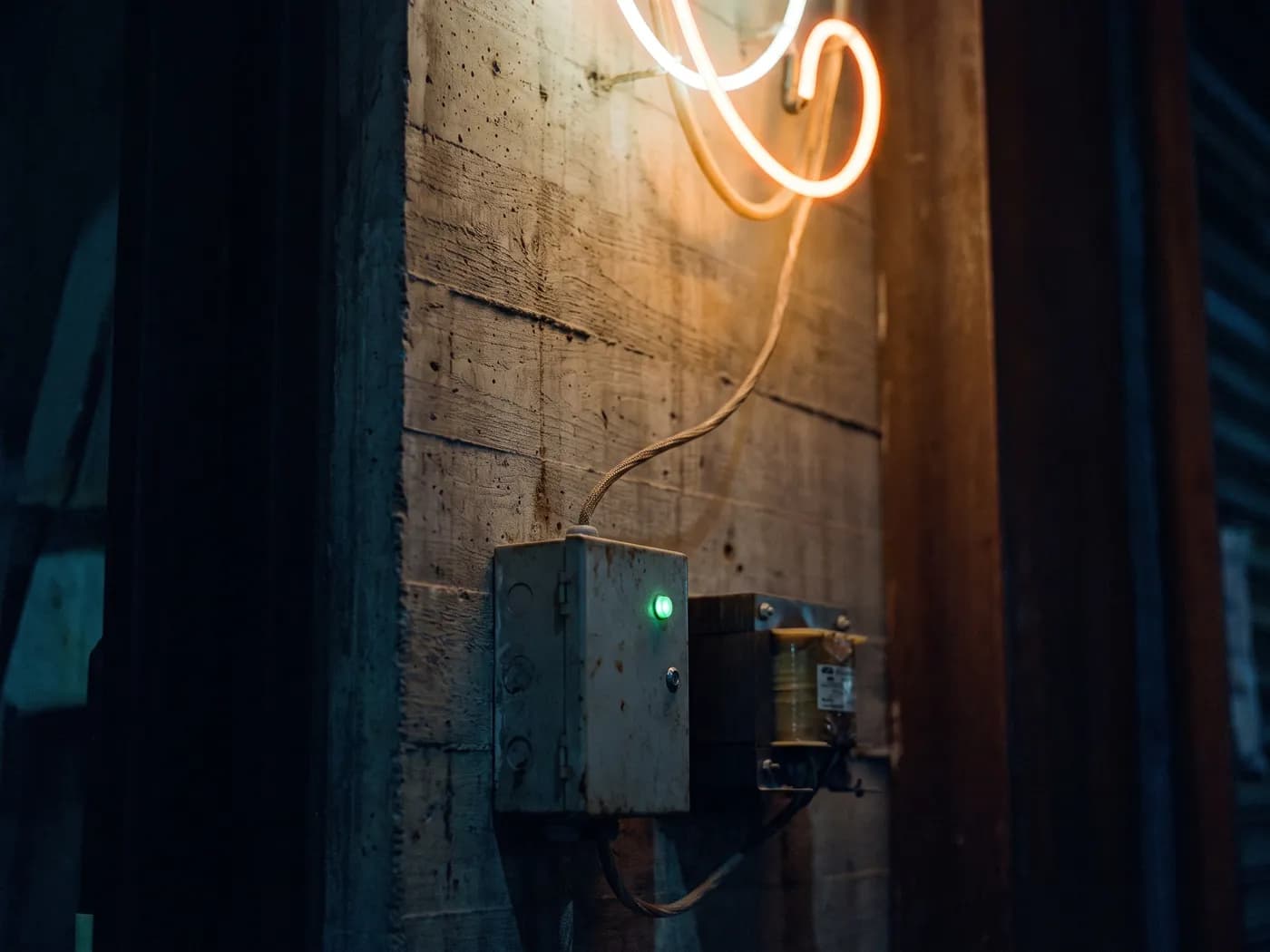 A small controller box with one green indicator lamp mounted beside a neon transformer on a concrete wall, a cable running up to the lit loop above