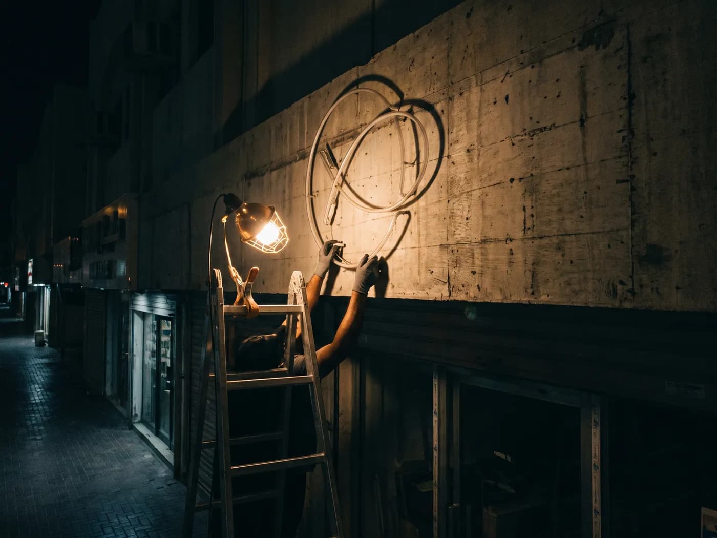 Two gloved hands on a ladder lifting a new, unlit neon loop into place above a shop door at night, one work lamp clipped to the ladder